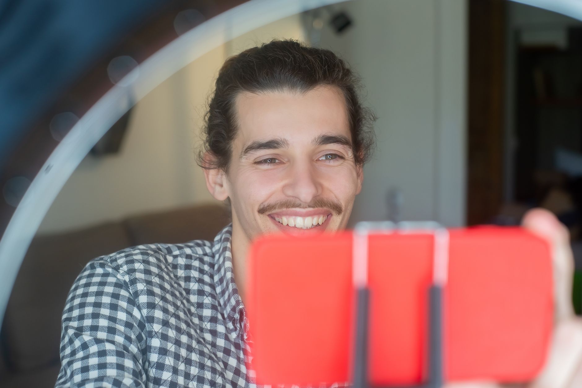 Un giovane sorridente si riprende con lo smartphone fissato a una luce ad anello, in una stanza di casa.